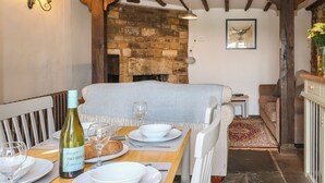 A dining area with a stone fireplace, wooden beams, a table set for two, and a sofa in the background. - Red Fawn Cottage (Moreton-in-Marsh)