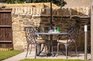 Un patio con un muro de piedra, cerca de madera y sillas y mesa de metal.
