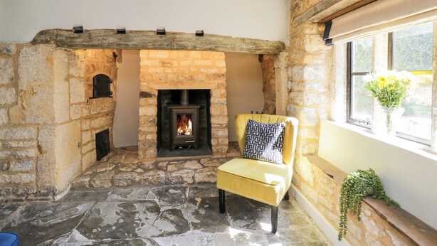 Una habitación con piso de piedra, una chimenea, un sillón amarillo con cojín estampado y una ventana con planta.