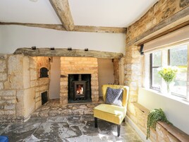 Una habitación con piso de piedra, una chimenea, un sillón amarillo con cojín estampado y una ventana con planta.