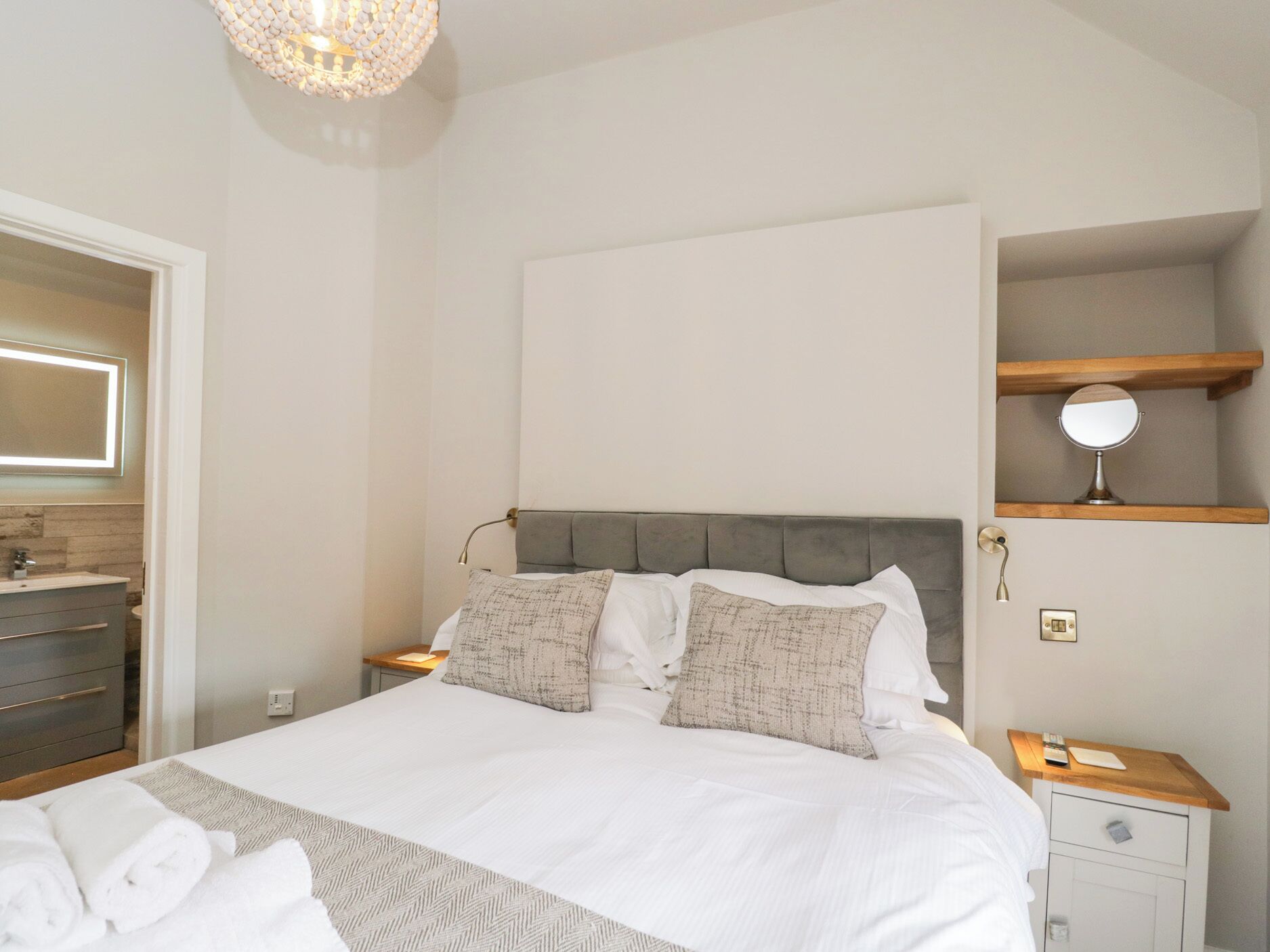 A neatly arranged bedroom with a large bed, bedside tables, a mirror, and a shelf with a lamp.
