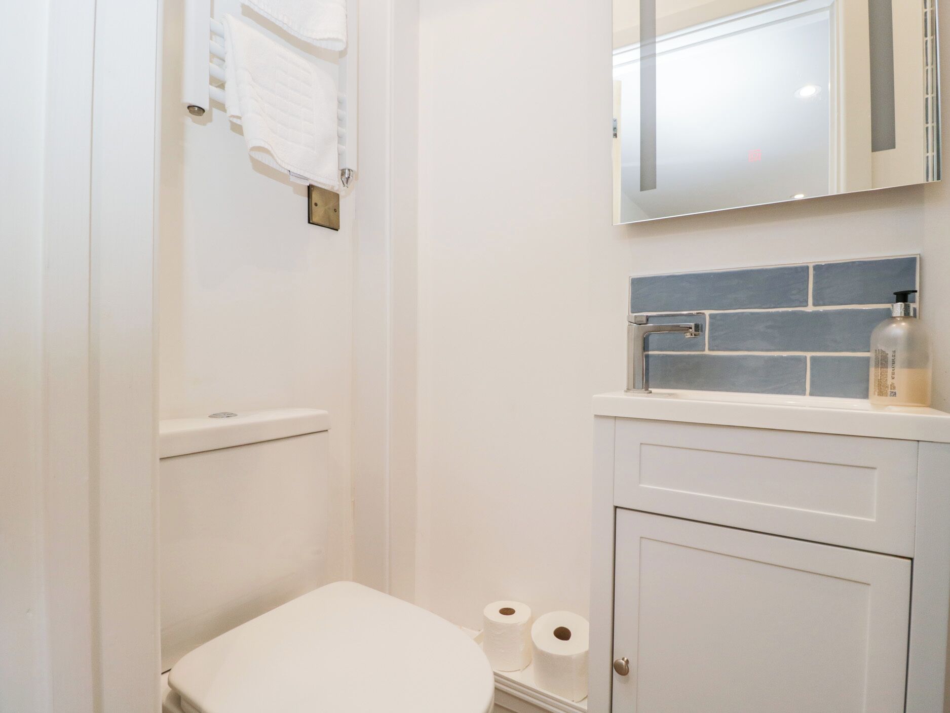 Baño pequeño y blanco con inodoro, lavabo y espejo. Hay dos rollos de papel higiénico en el suelo.