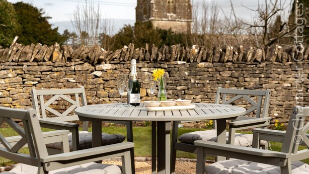 Une table ronde avec quatre chaises, une bouteille de champagne et un vase rempli de fleurs, le tout placé devant un mur de pierre avec une tour en arrière-plan.