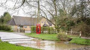 Un paysage rural avec une cabine téléphonique rouge traditionnelle, une maison au toit en pente et un petit ruisseau.