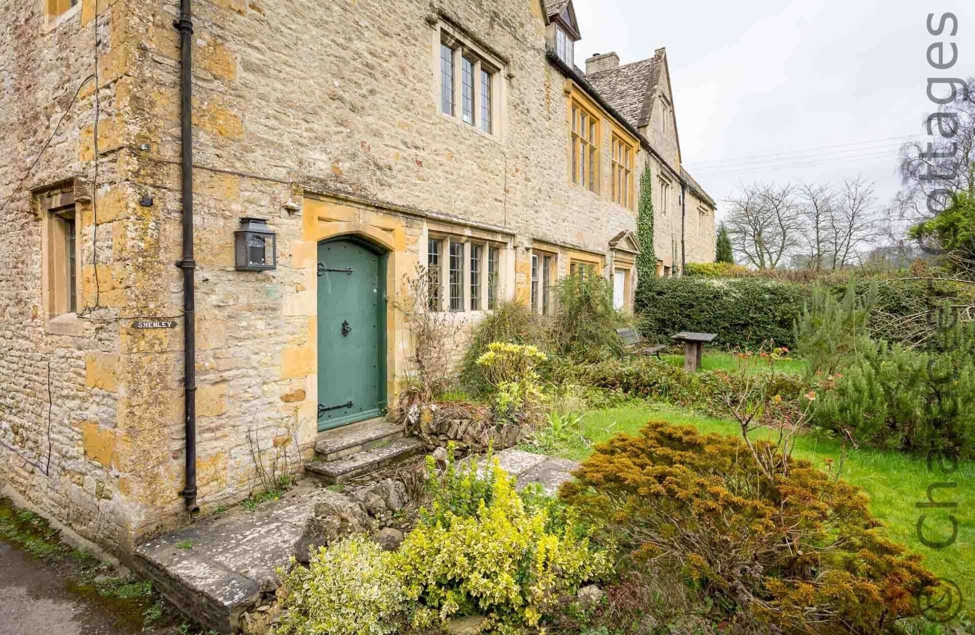 Un bâtiment en pierre avec une porte verte, un petit jardin avec différentes plantes et une enseigne indiquant « SENLEY ».