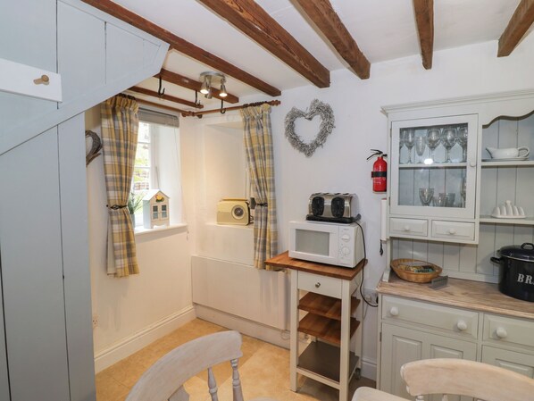 A kitchen with a white microwave, a toaster, and a small table with a basket. There is a window with curtains and a heart-shaped wreath on the wall. - Jackdaw Cottage (Moreton-in-Marsh)