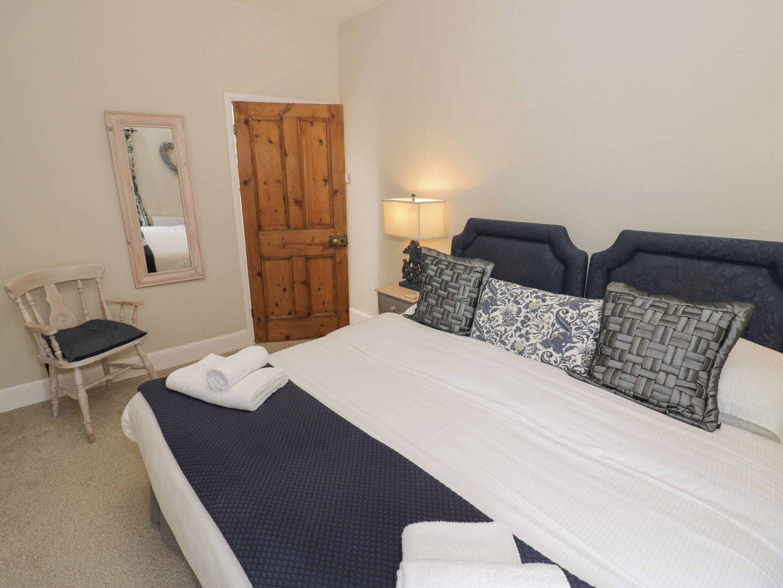 A neatly made bed with a dark blue bedspread, a wooden door, a chair with a cushion, and a wall-mounted mirror.