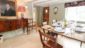 A dining area with a wooden table set for four, surrounded by wooden chairs. A vintage lamp and a framed portrait are on the wall. - Holly Cottage (Witney)