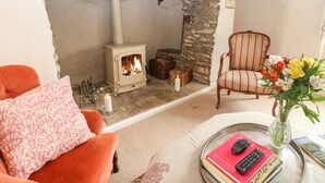 A cozy living room with a stone fireplace, a plush orange armchair, a striped armchair, and a table with books and a vase of flowers. - Holly Cottage (Witney)