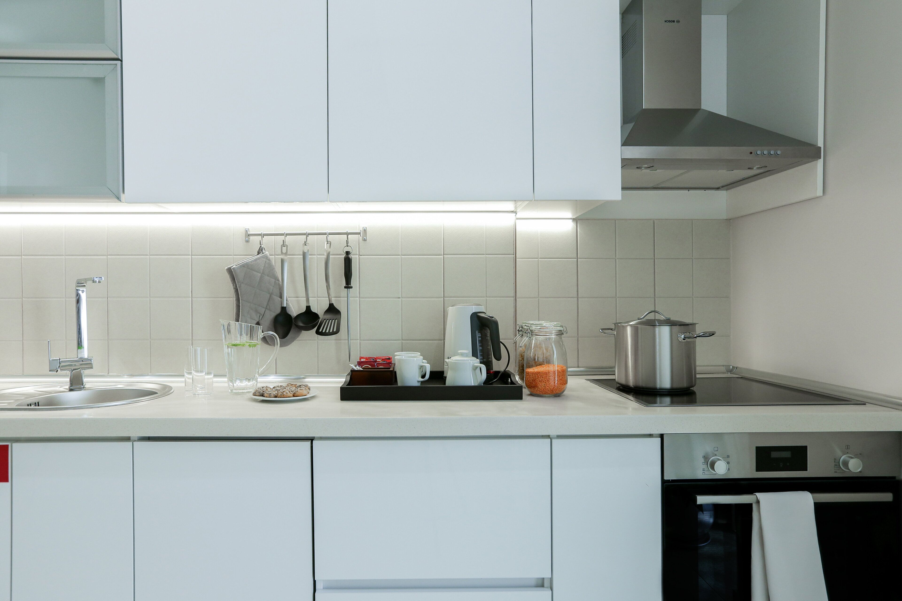 Uma cozinha moderna com armários brancos, um exaustor de aço inoxidável, uma máquina de lavar louça e uma bancada com diversos utensílios de cozinha.