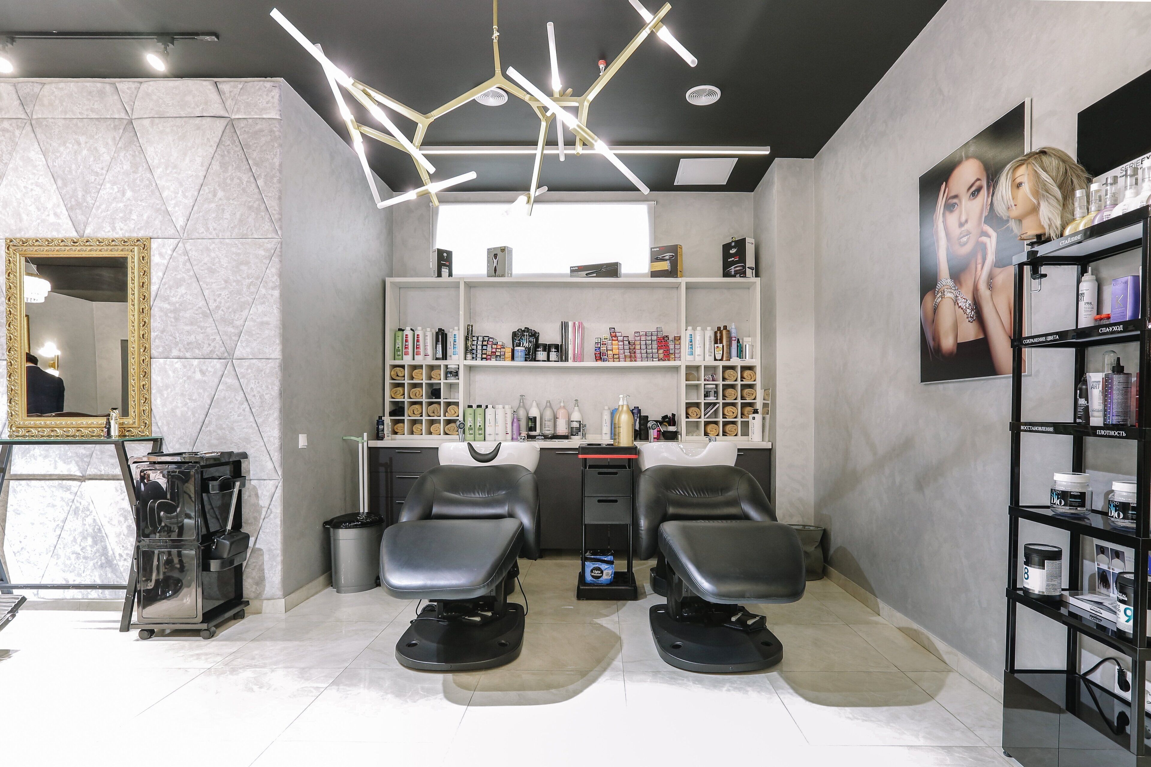 Un salone di bellezza con due poltrone nere, uno specchio con cornice dorata, scaffali con prodotti per capelli e un moderno lampadario.