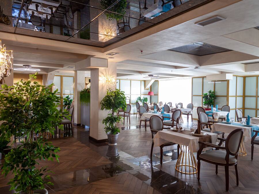 Een ruime eethoek met meerdere tafels en stoelen, potplanten en een hoog plafond met zichtbare balken.