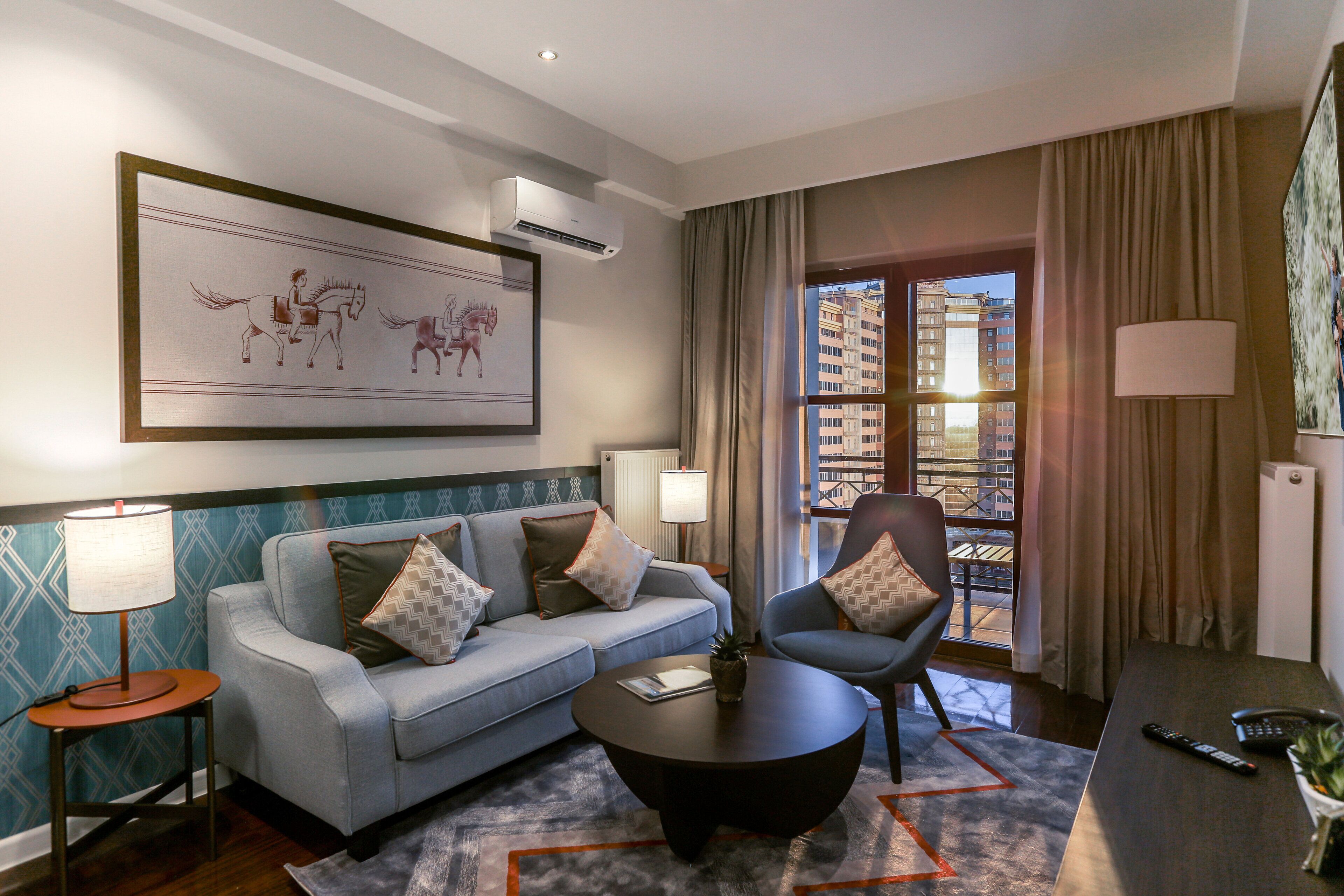 Ein Hotelzimmer mit Sofa, Sesseln, einem Couchtisch, einem Gemälde und Blick auf die Stadt.