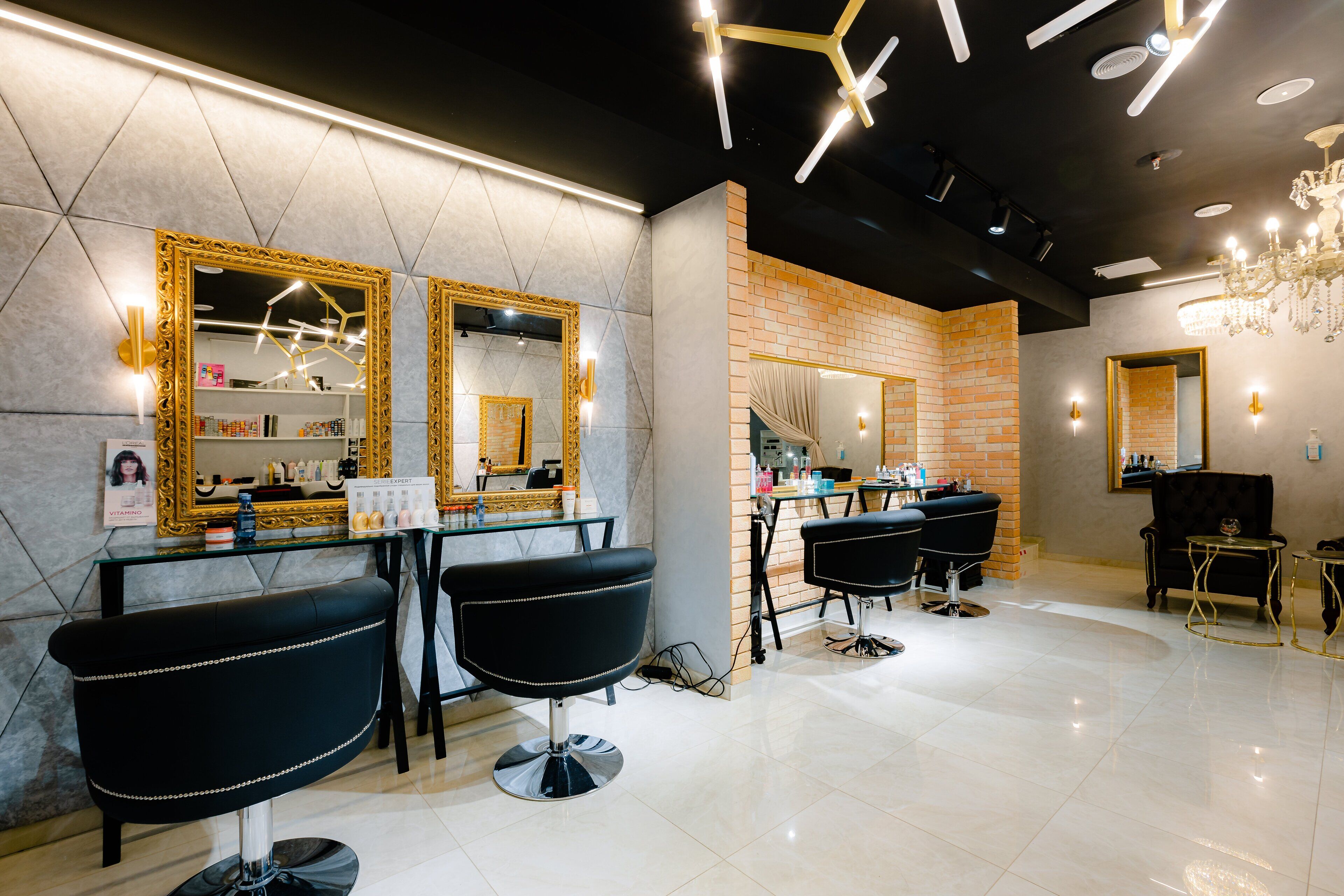 A modern salon with three black salon chairs, a mirror with a gold frame, and a chandelier.