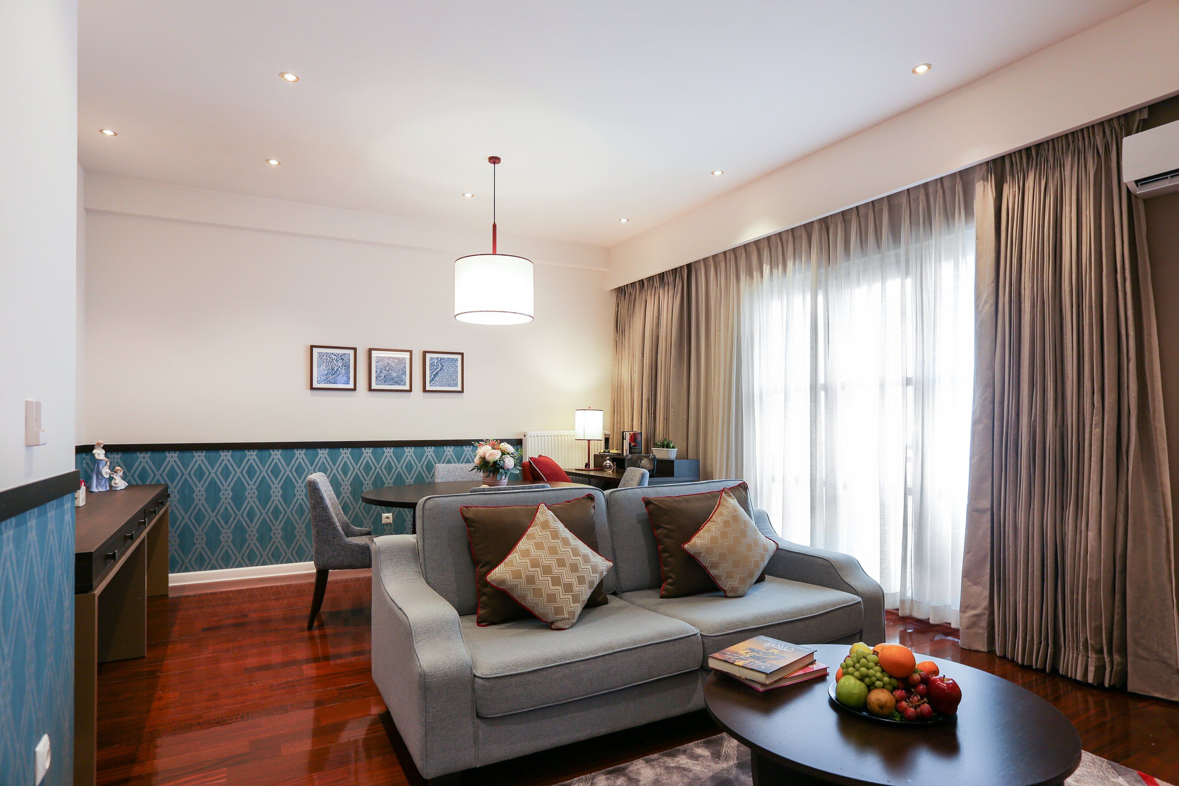 Uma sala de estar moderna com um sofá cinzento, uma mesa de centro com frutas, uma área de jantar com mesa e cadeiras, e uma janela grande com cortinas.