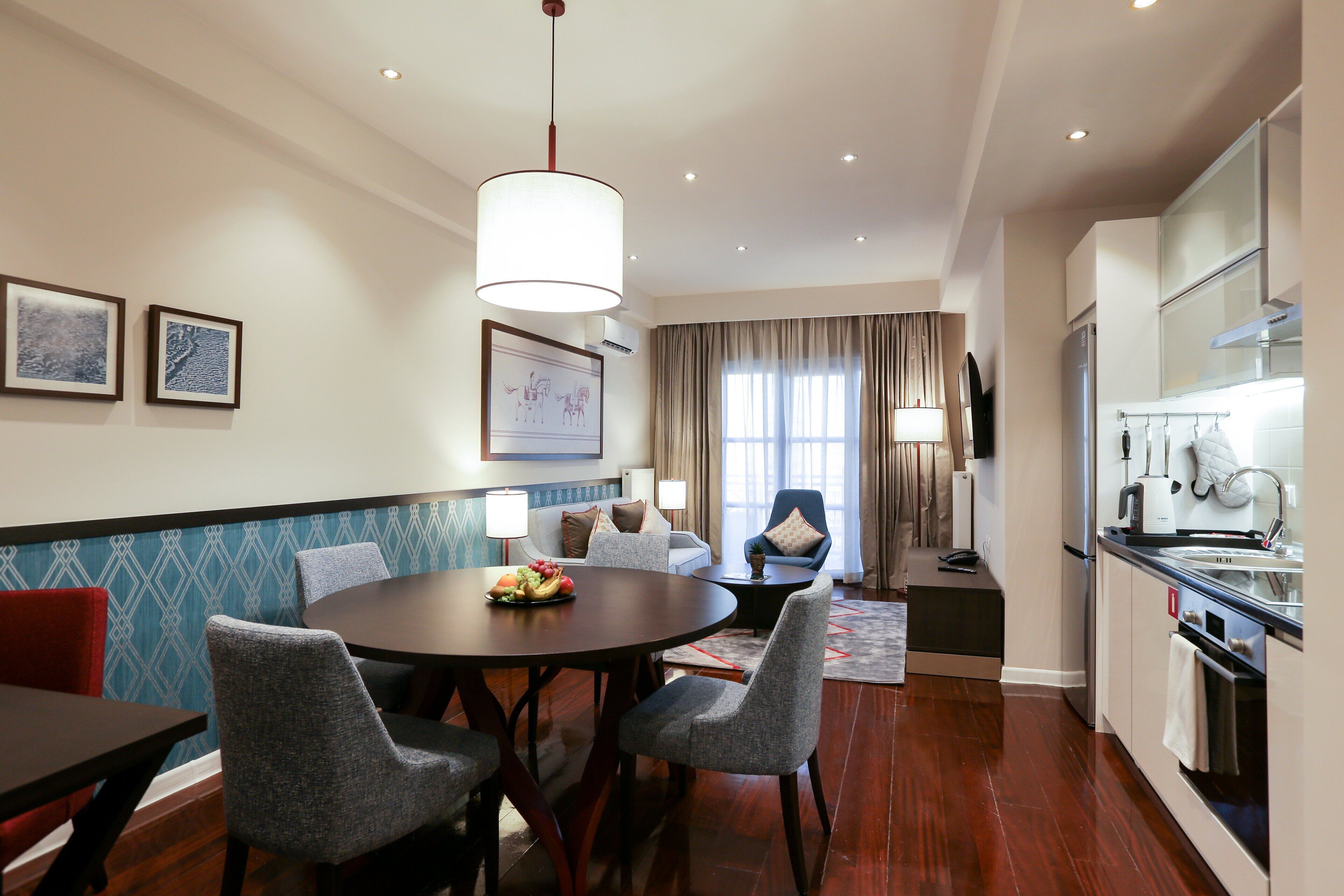 A modern hotel room with a dining area, a living area with a sofa and armchairs, a kitchen with a refrigerator and stove, and a large window with curtains.