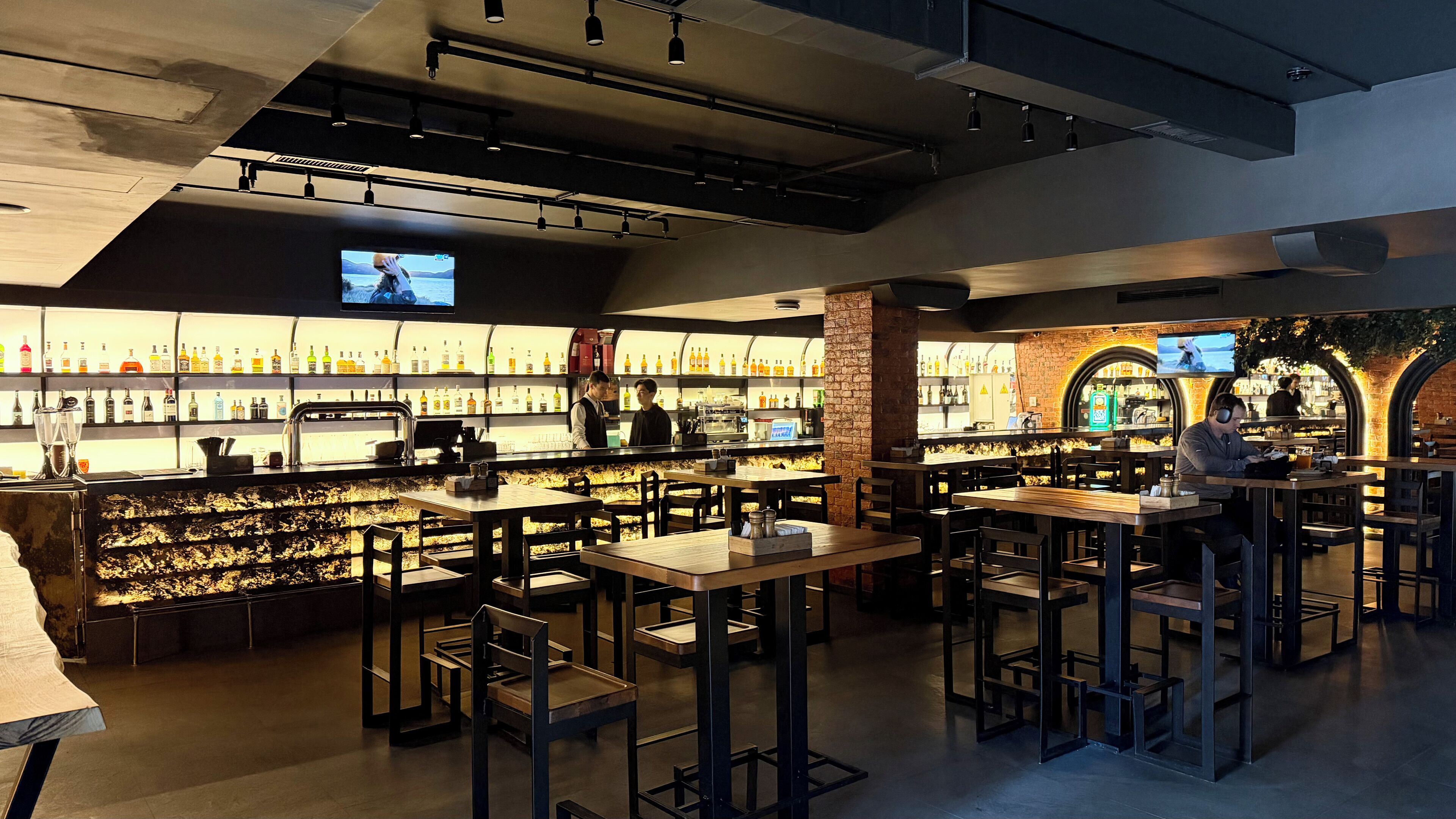 A bar area with multiple seating options, a television screen, and a variety of bottles on shelves.