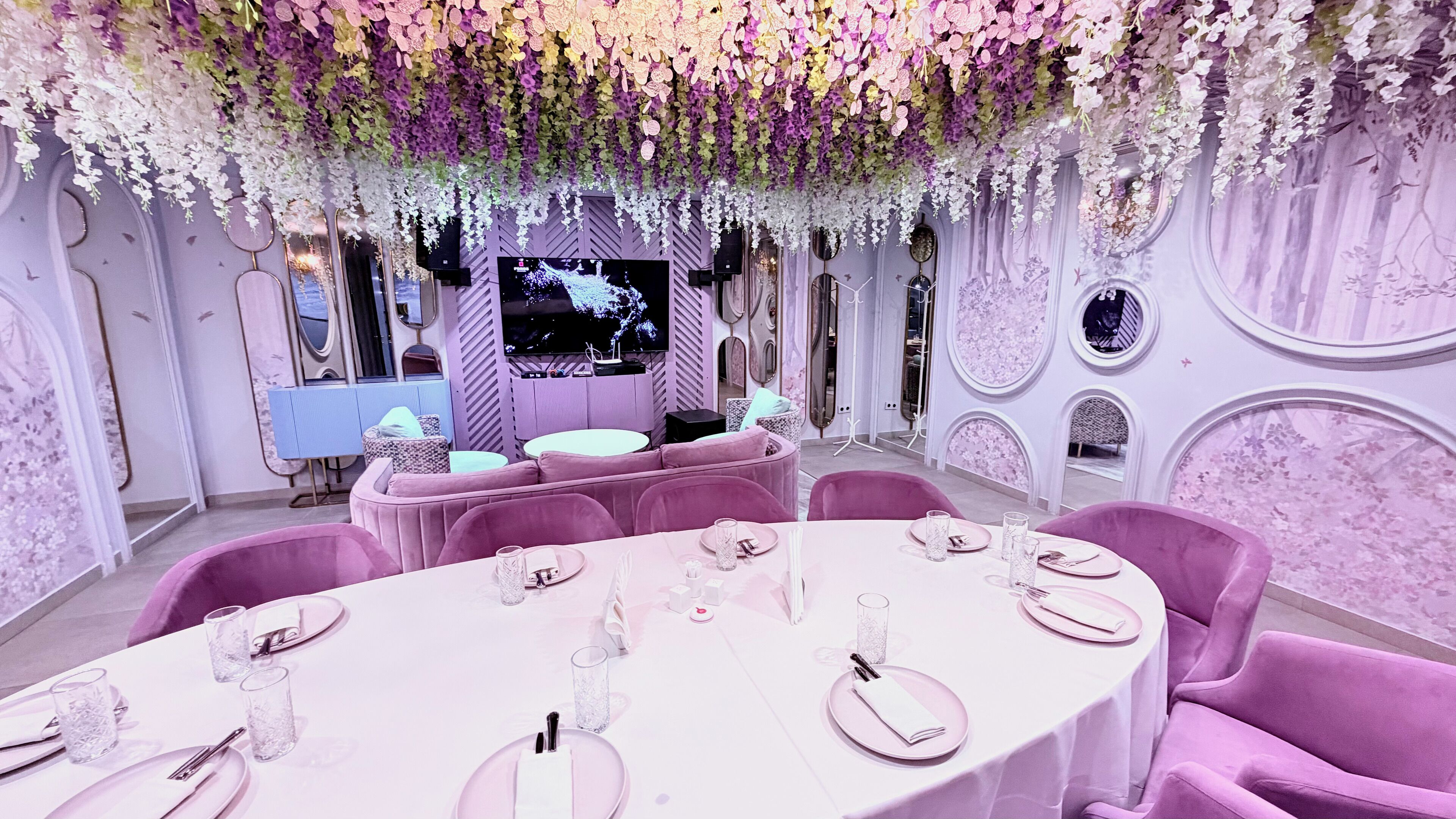 A luxurious dining area with round tables, pink chairs, and a ceiling adorned with hanging flowers.