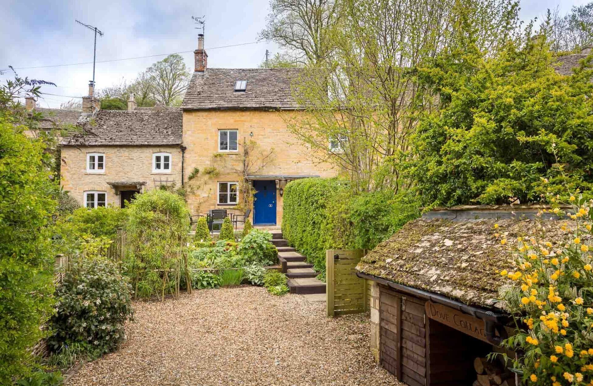 A stone house with a blue door, surrounded by lush greenery and a gravel path leading to the entrance.