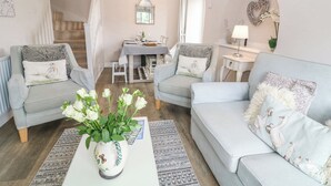 A cozy living room with two light grey sofas, a white coffee table with a vase of flowers, a dining area with a table set for four, and a staircase leading to an upper floor. - Honeysuckle Cottage (Banbury)