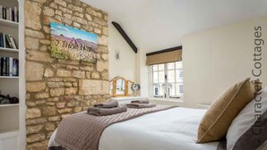 A bedroom with a stone wall, a bed with a beige and gray duvet, a window with a view, and a bookshelf with books. - Lanes Cottage (Chipping Campden)