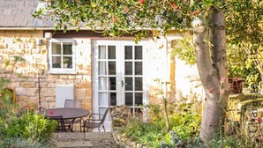 A stone building with a patio area featuring a table and chairs, surrounded by lush greenery and a stone pathway. - Lanes Cottage (Chipping Campden)