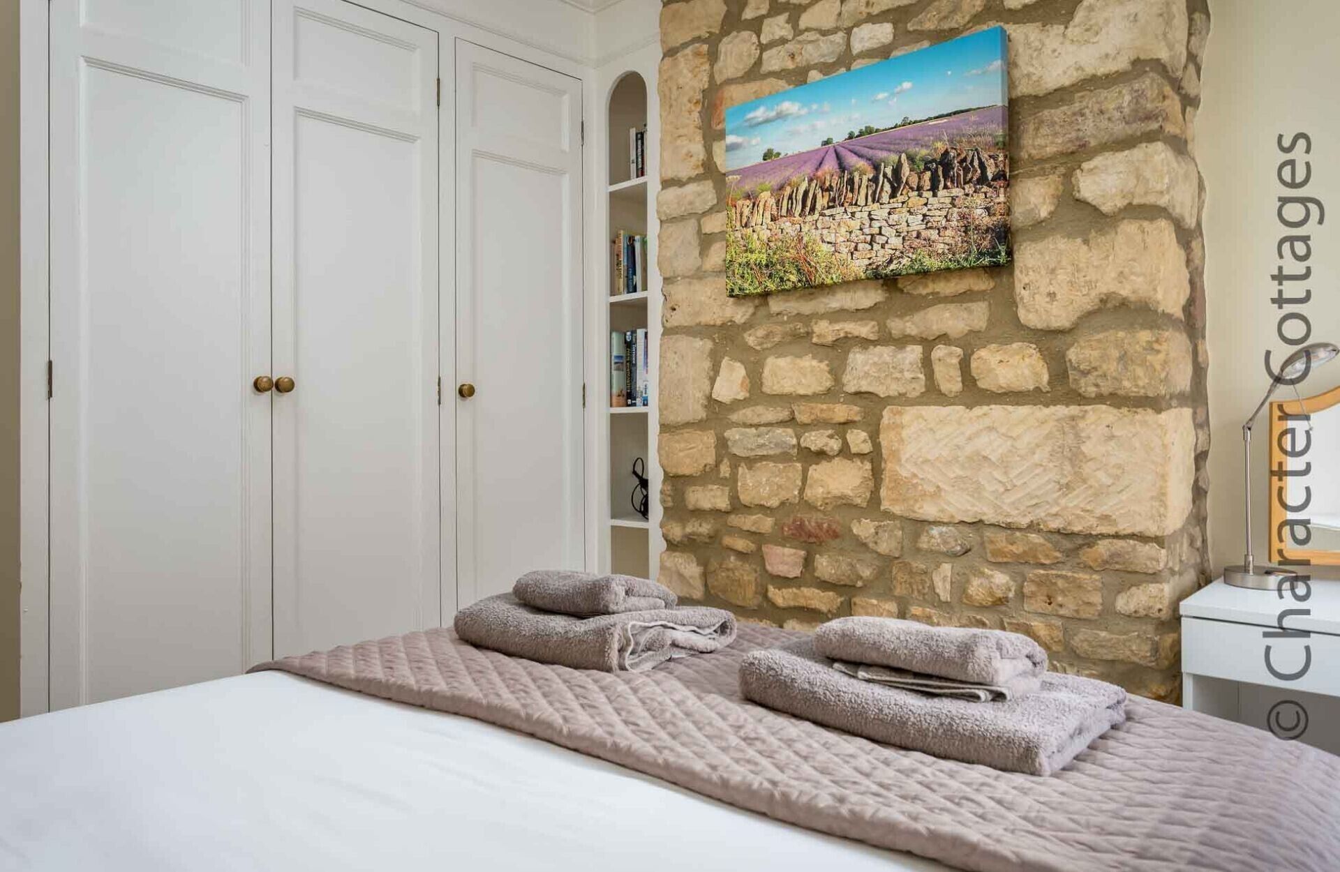 Une chambre avec un mur en pierre, un lit avec une couette et des serviettes, une bibliothèque et un tableau.