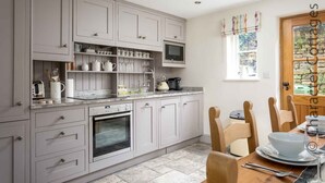 A modern kitchen with grey cabinets, a built-in oven, a microwave, and a coffee machine. There is a dining table with chairs and a window with curtains.