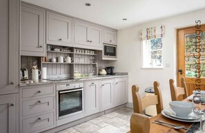 A modern kitchen with grey cabinets, a built-in oven, a microwave, and a coffee machine. There is a dining table with chairs and a window with curtains. - Willow Vale (Broadway)