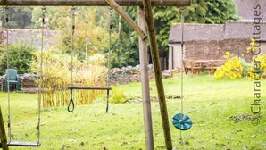A wooden swing set in a garden with a grassy lawn, a bench, and a small shed in the background.
