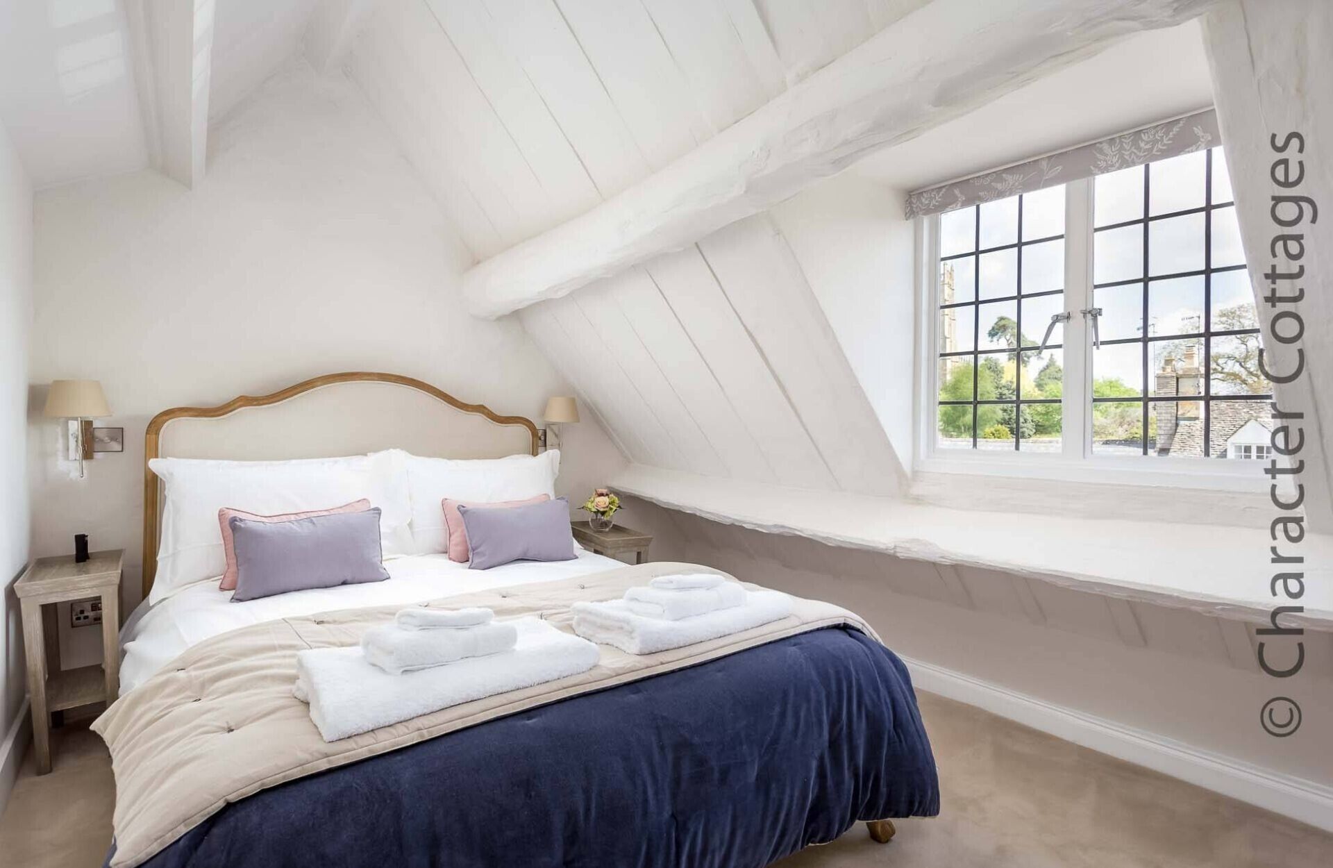 Un dormitorio con techo inclinado, un ventanal con vistas a la vegetación, una cama con ropa blanca y varias almohadas, y una mesita de noche con una lámpara.