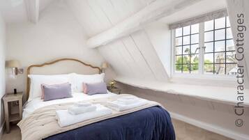 Un dormitorio con techo inclinado, un ventanal con vistas a la vegetación, una cama con ropa blanca y varias almohadas, y una mesita de noche con una lámpara.