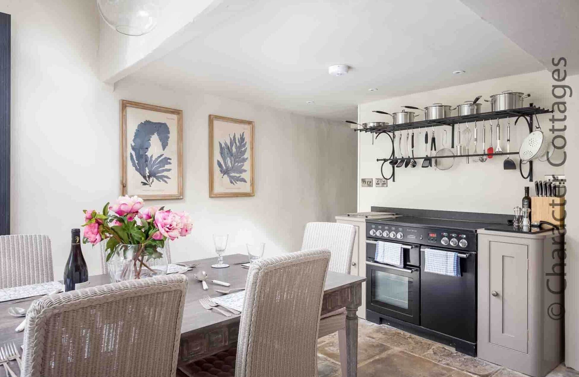 Un comedor con sillas de mimbre y una mesa preparada para dos, una cocina con estufa y horno, y un soporte de utensilios en la pared.