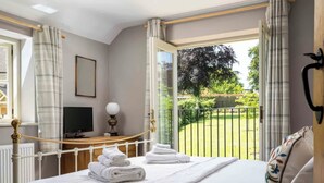 Een net opgemaakt bed met witte lakens en handdoeken, een nachtkastje met een lamp, een televisie en uitzicht op een groene tuin door een open raam.