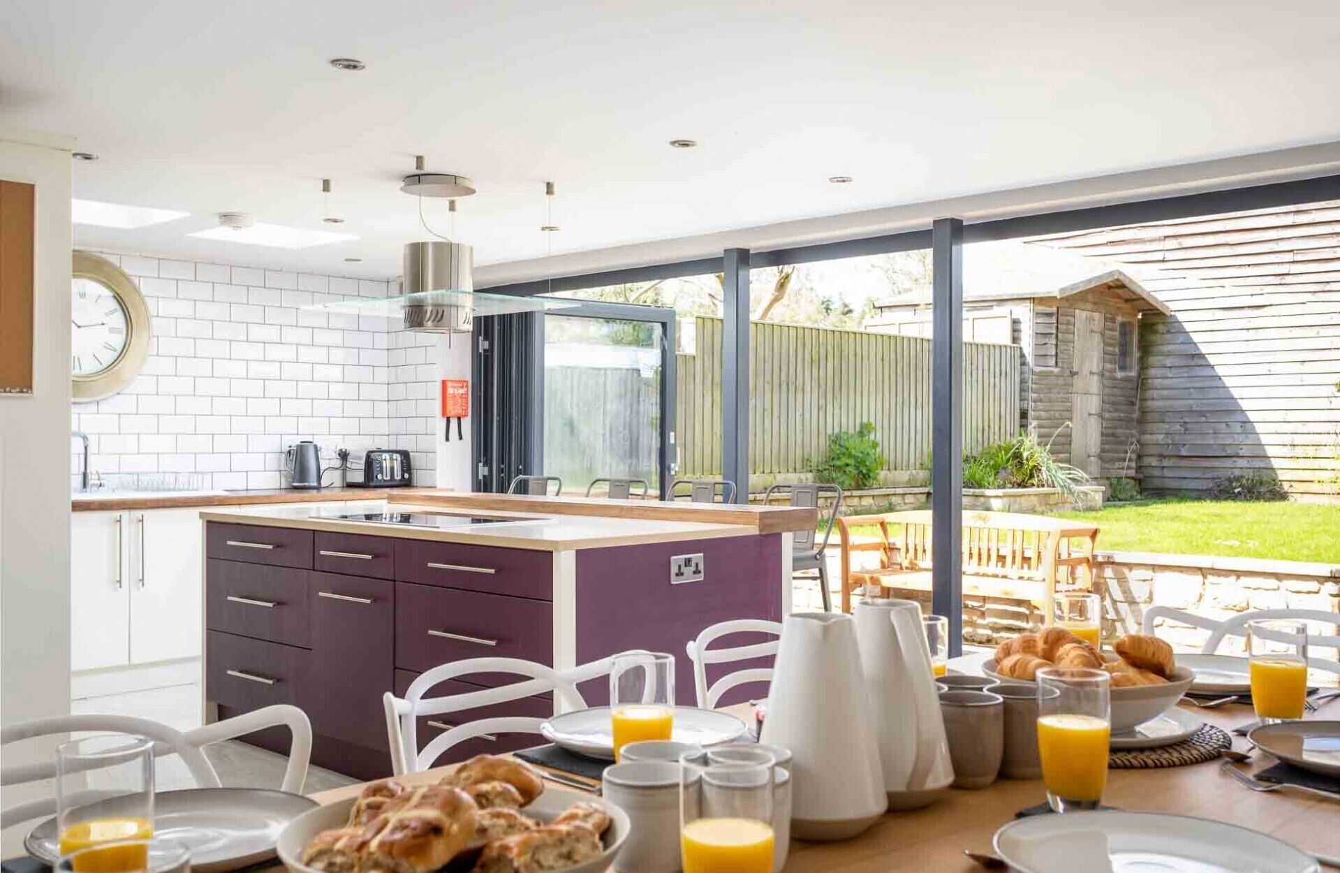 A modern kitchen with a large island, white tiled backsplash, and a view of the backyard through large windows.