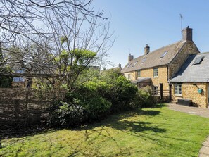 A stone house with a sloped roof, a visible chimney, and a small garden area with grass and a wooden fence.
