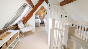 Ein gemütliches Schlafzimmer im Dachgeschoss mit freiliegenden Holzbalken, einer weißen Treppe, einer Holzbank und Blick in den Wohnbereich.