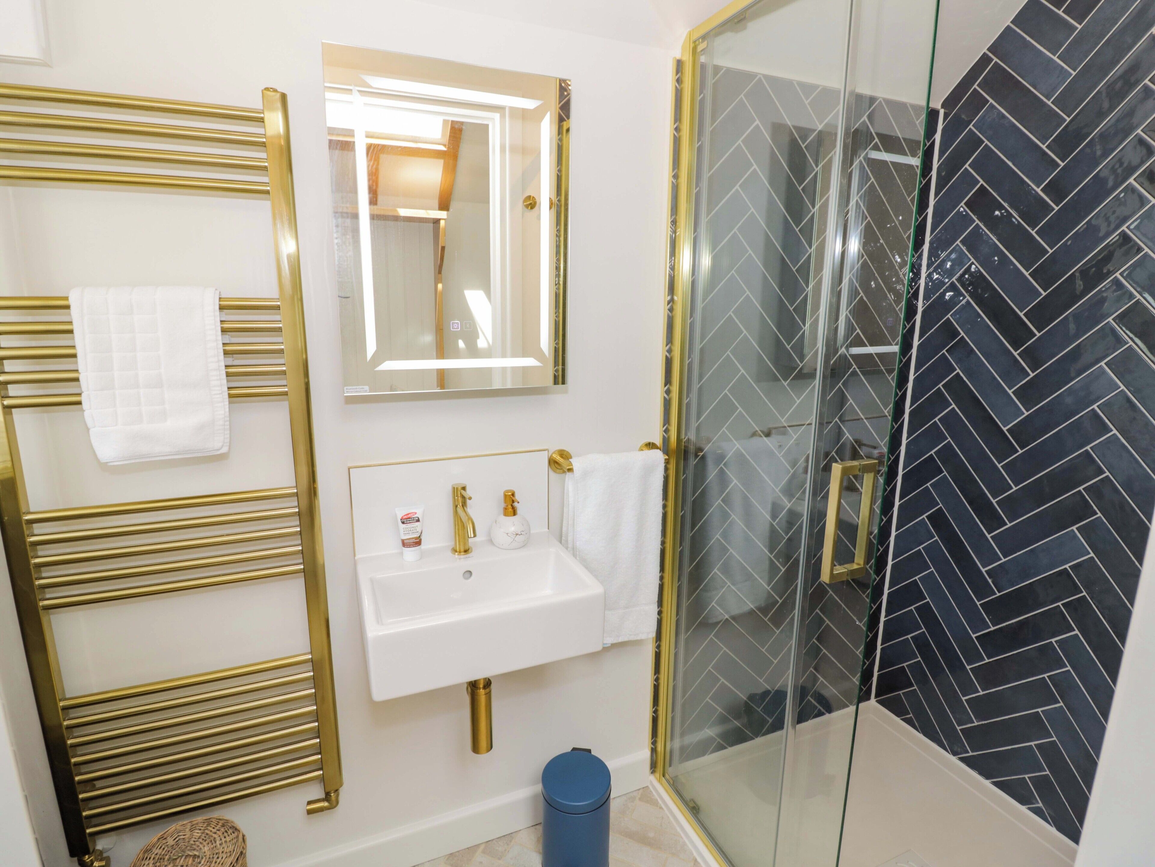 Un baño con un espejo de marco dorado, un lavamanos blanco con detalles dorados y una ducha con salpicadero de azulejos de espiga en blanco y negro.