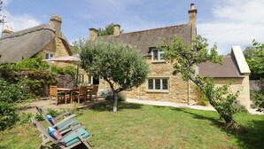 Une maison en pierre avec un toit de chaume, une terrasse avec des meubles en bois et un petit arbre dans la cour.