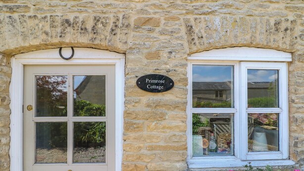 A stone cottage with a white door and window, a sign reading "Primrose Cottage," and a flower box with red flowers. - Primrose Cottage (Cheltenham)