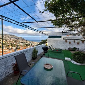 Eine Terrasse mit Glastisch, Stühlen, einem Grill und Blick auf eine Küstenstadt.