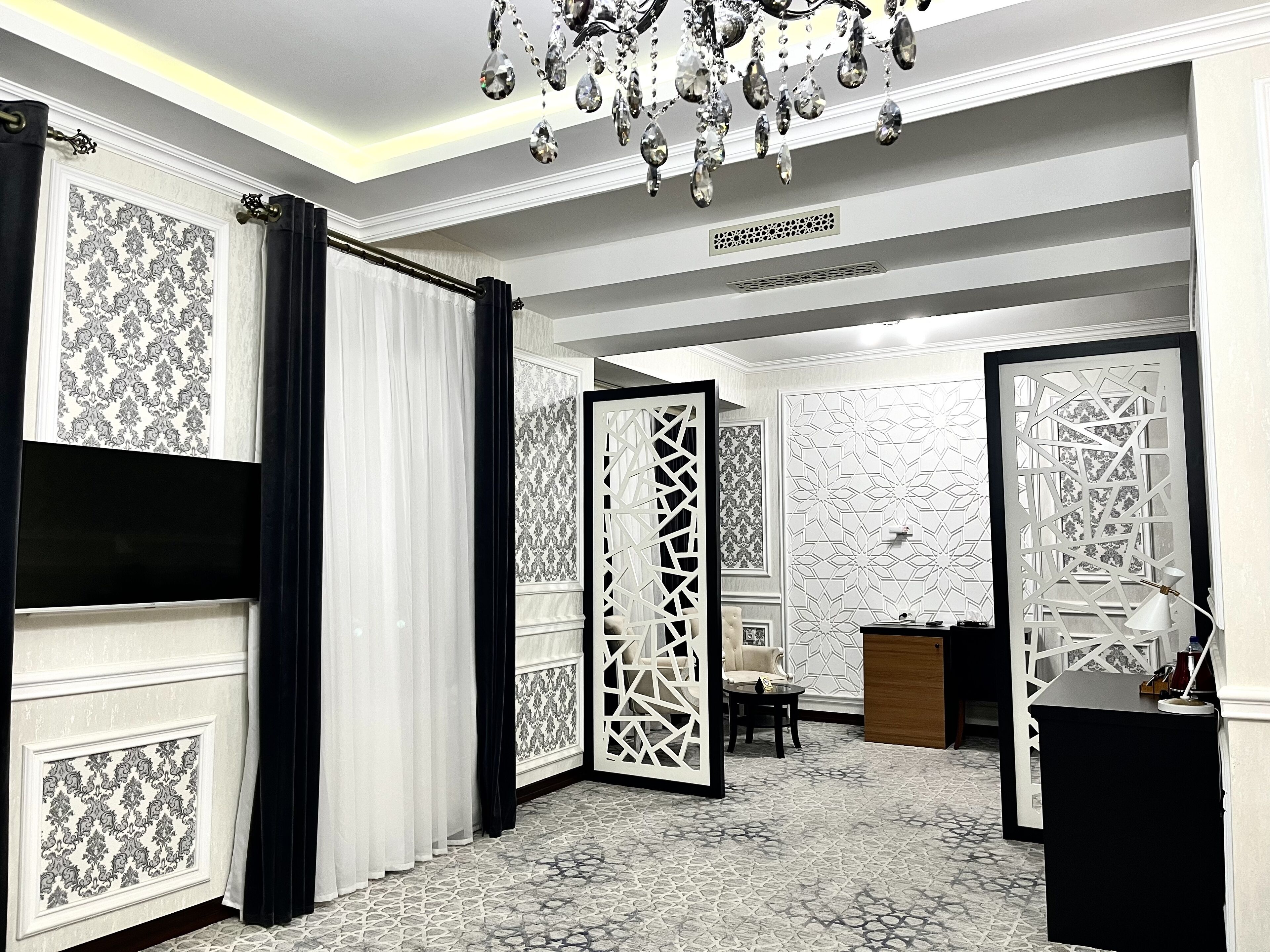 A room with a patterned wallpaper, a chandelier, a white partition, and a desk with a lamp.