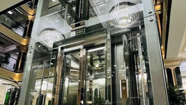 A modern elevator with glass doors and a metallic interior, surrounded by marble flooring and a high ceiling with recessed lighting. - Garden Plaza Bukhara (Bukhara)