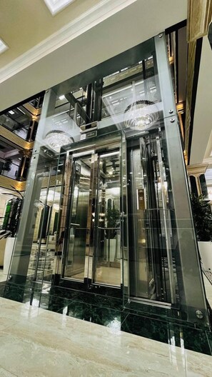 Een moderne lift met glazen deuren en een metalen interieur, omringd door een marmeren vloer en een hoog plafond met inbouwverlichting.