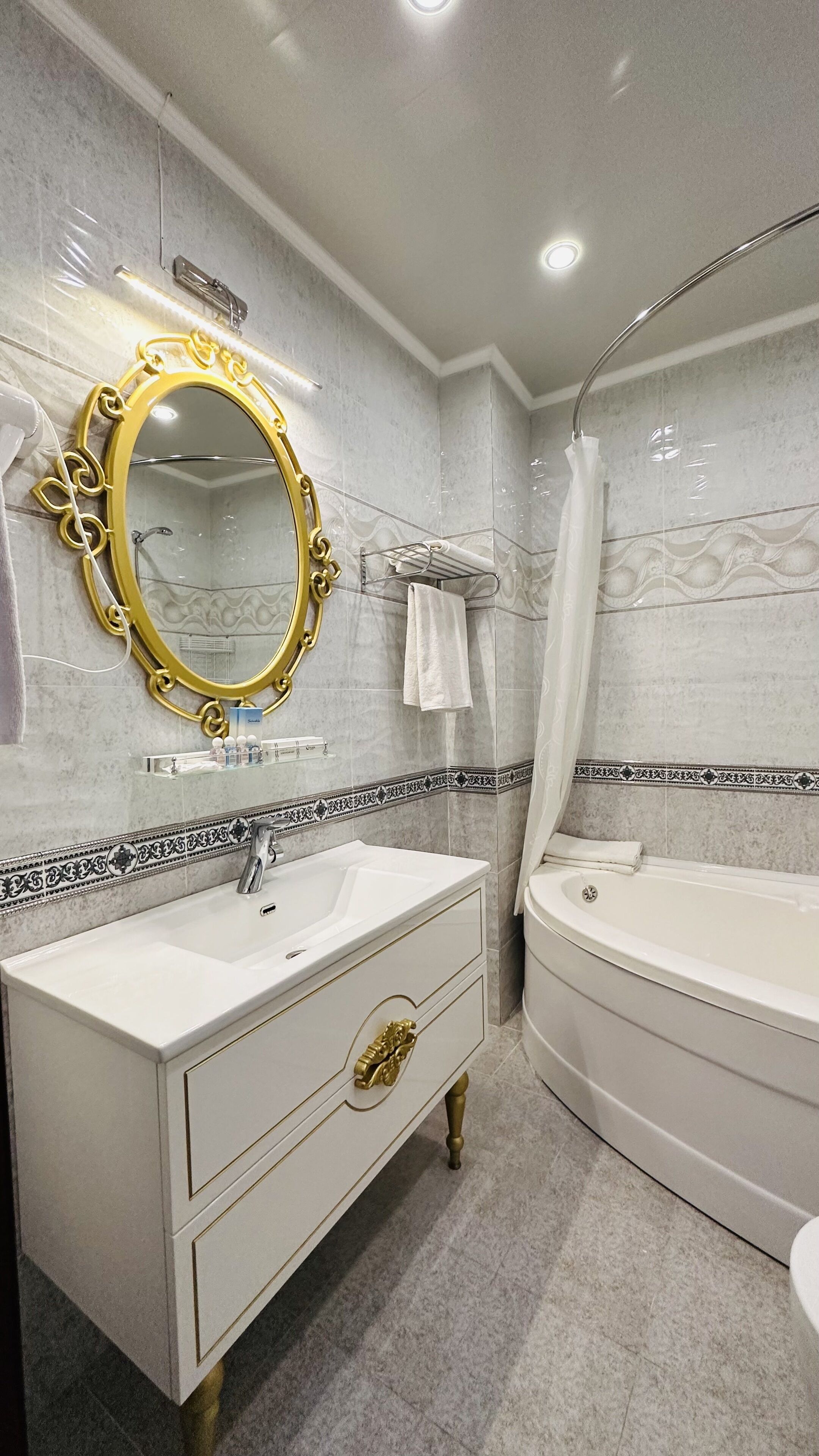 Une salle de bain avec un meuble-lavabo blanc, un miroir encadré d’or, une baignoire et un rideau de douche.
