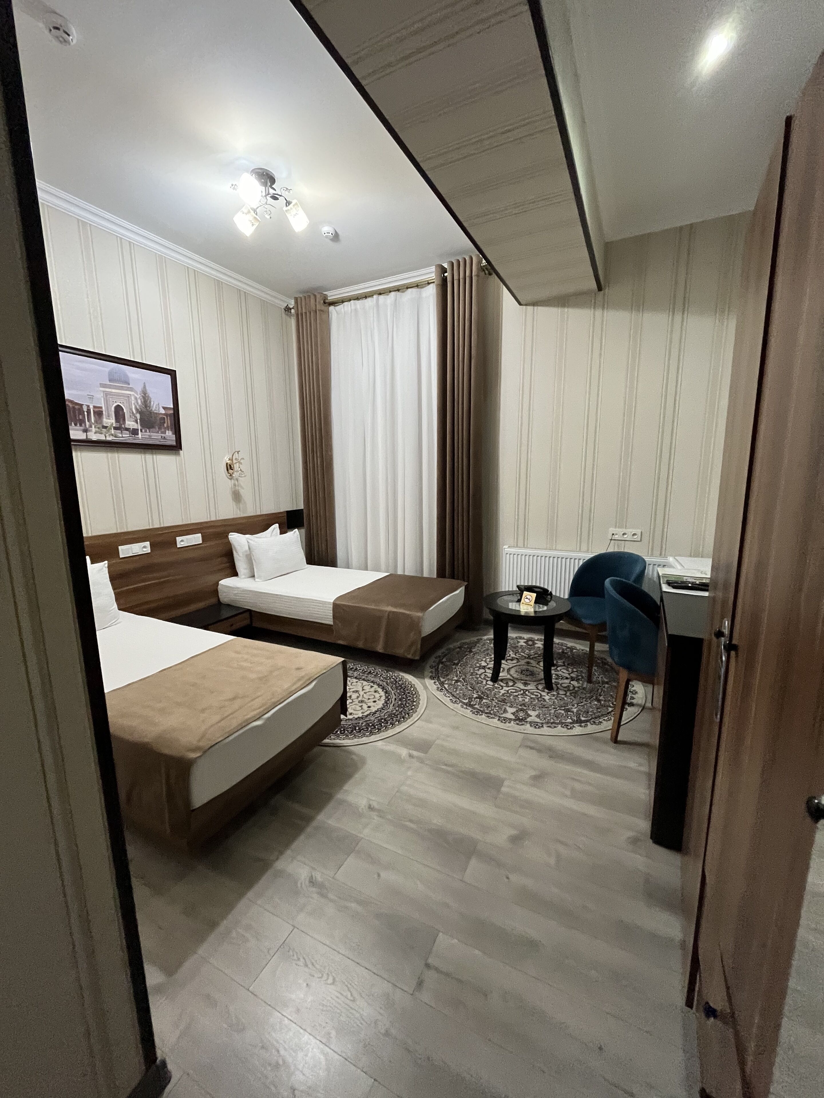 Une chambre d’hôtel avec deux lits, un bureau, une chaise, une petite table, un tapis rond et un tableau au mur.