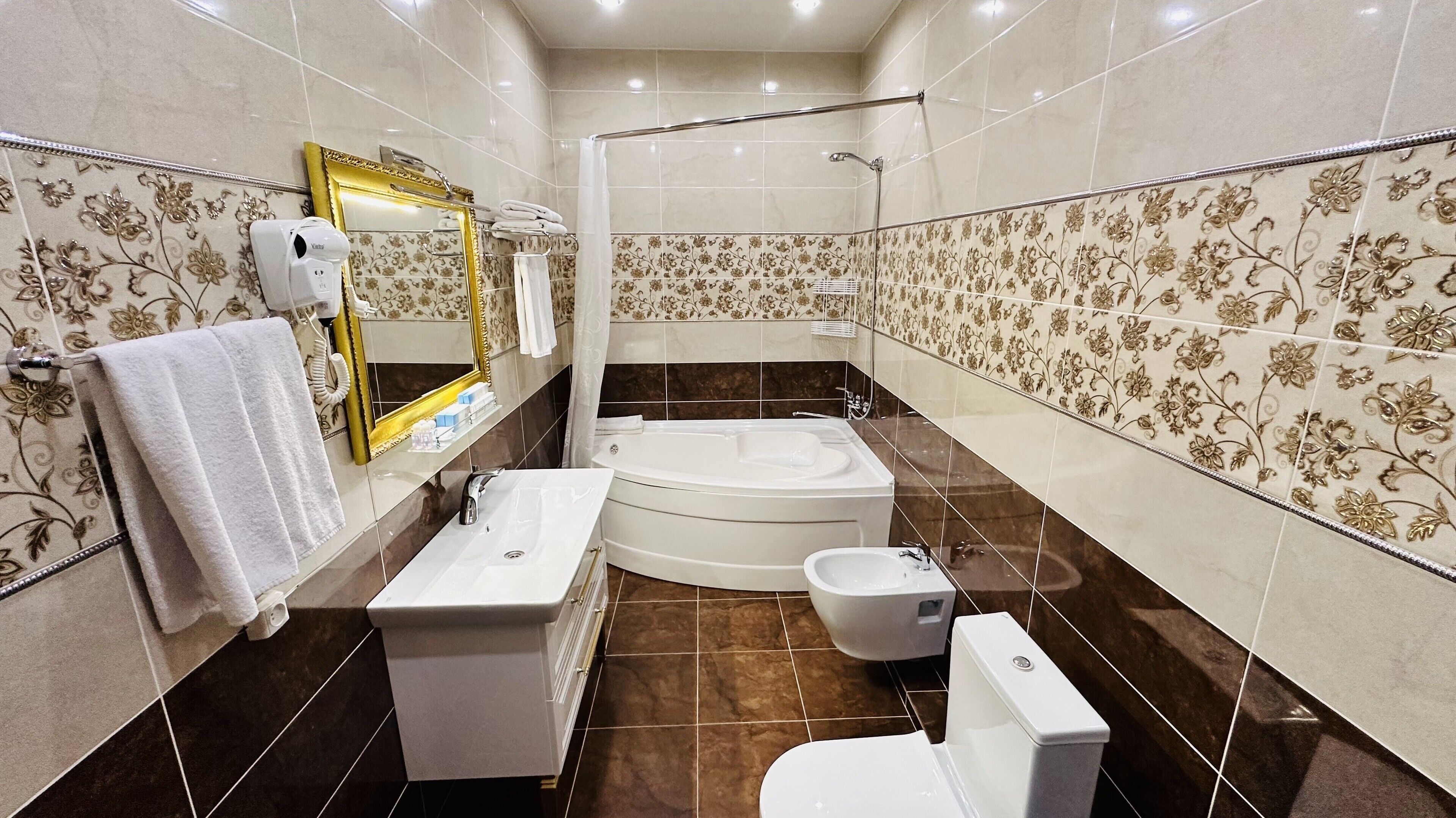 Casa de banho com banheira, sanita e lavatório, apresentando padrões de azulejos florais e um espelho com moldura dourada.