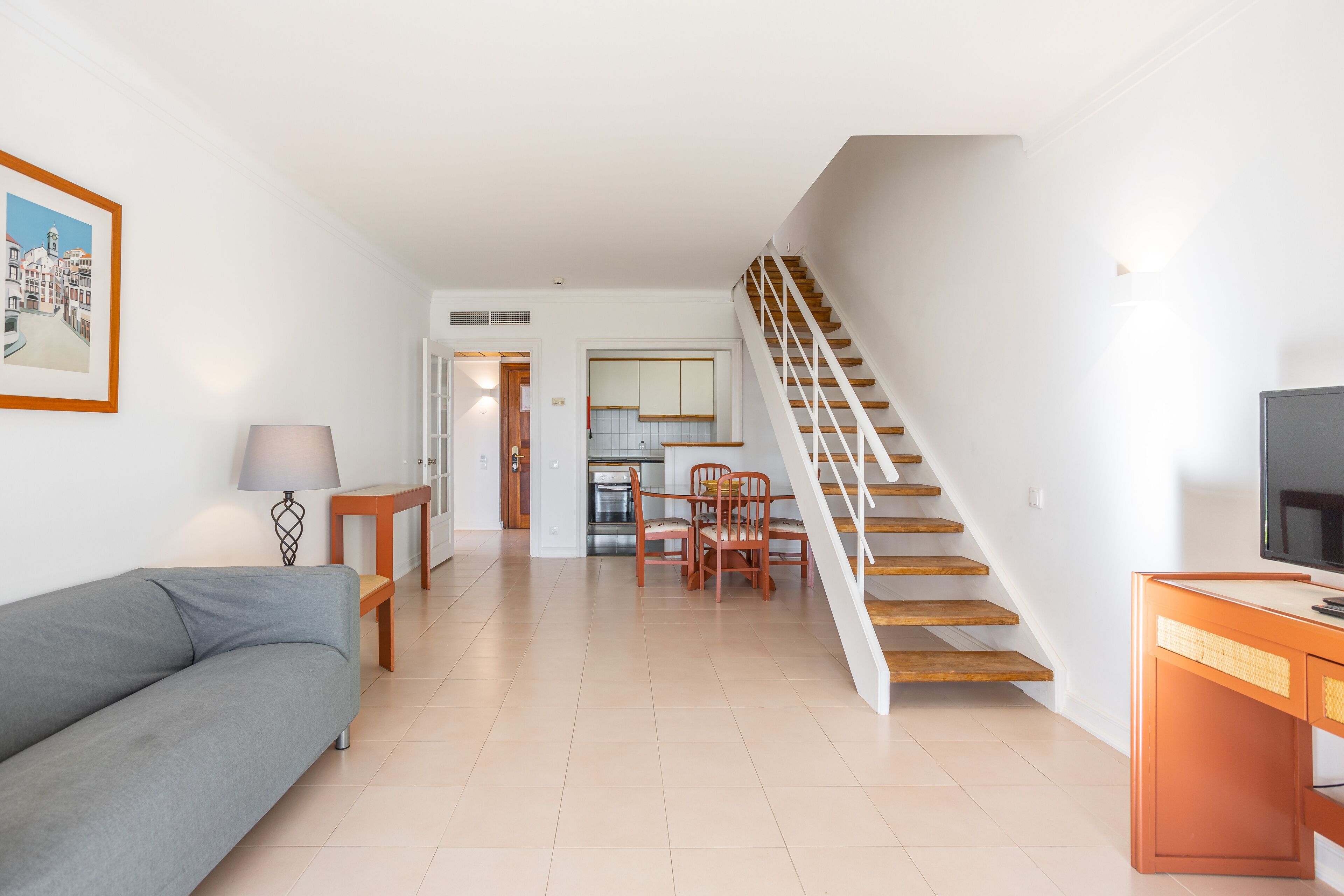 A modern living room with a sofa, a dining table with chairs, a staircase, and a kitchen area in the background.