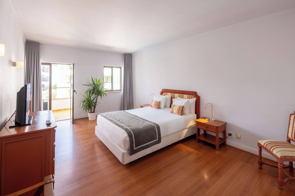 A hotel room with a large bed, bedside table, TV, and a potted plant.