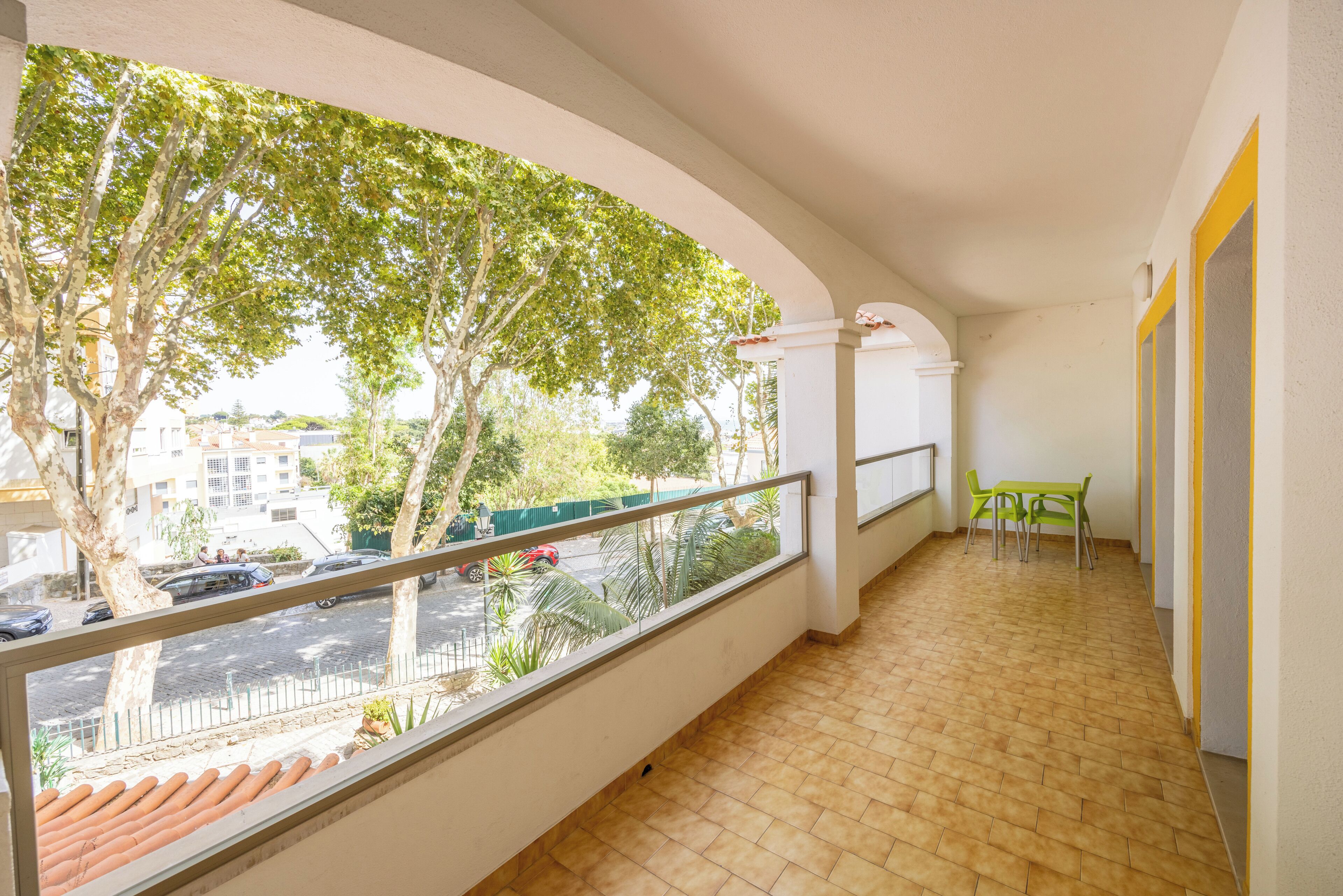 A balcony with a tiled floor, a view of a street with cars, and a green table with chairs.